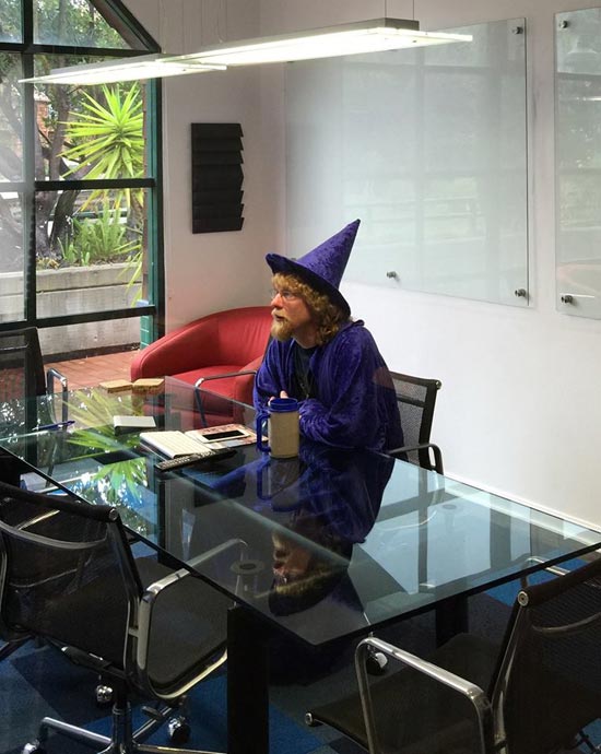 Wearing a Halloween costume of a purple wizard, sitting at a glass table in a conference room.