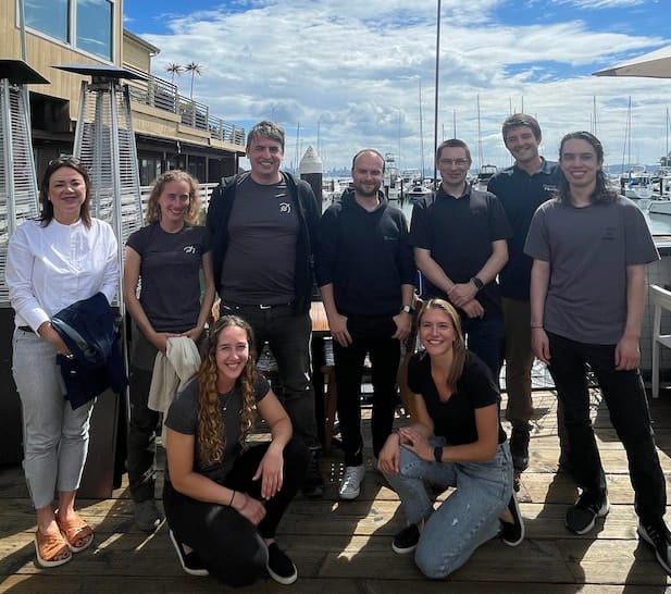 Nine-person team photo on a dock with sailboats in the background.