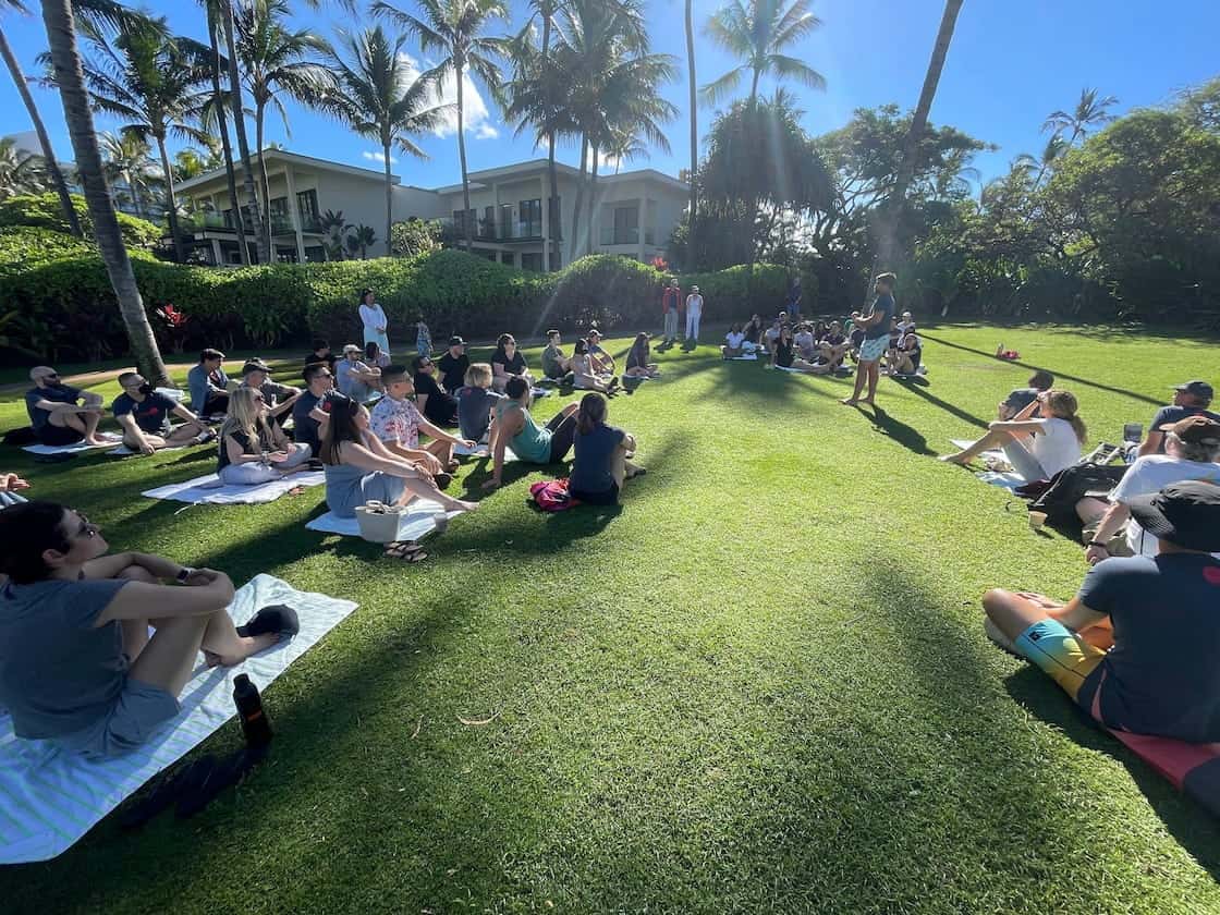 Large group sitting on a lawn under palm trees for a meeting.