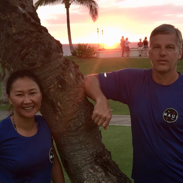 Couple leaning against a palm tree in front of a Hawaiian sunset.