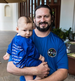 Bearded smiling man in Maui t-shirt holding a child.